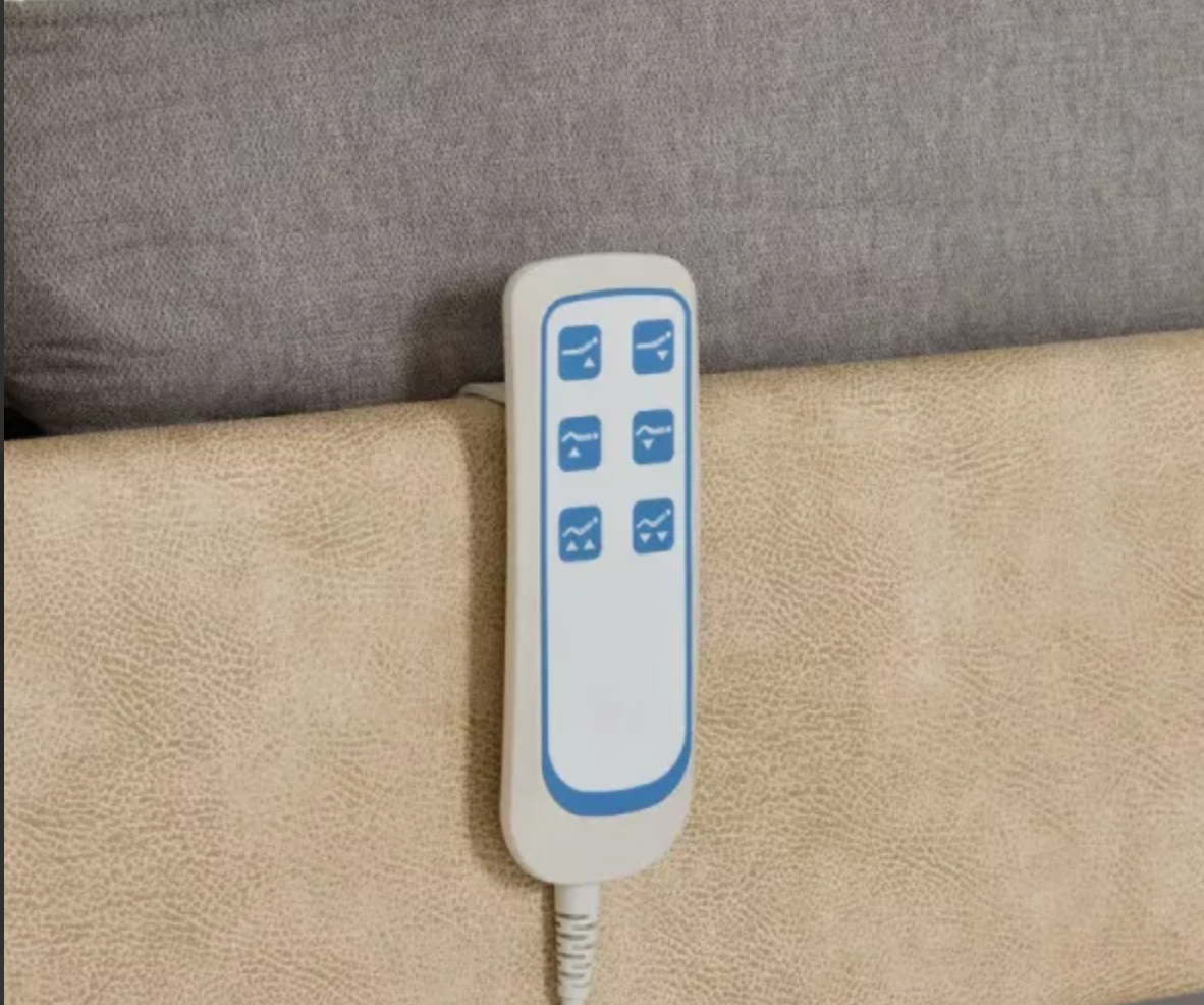 A close-up of an electric massage controller plug attached to a beige upholstered headboard or sofa, with a gray fabric background above.