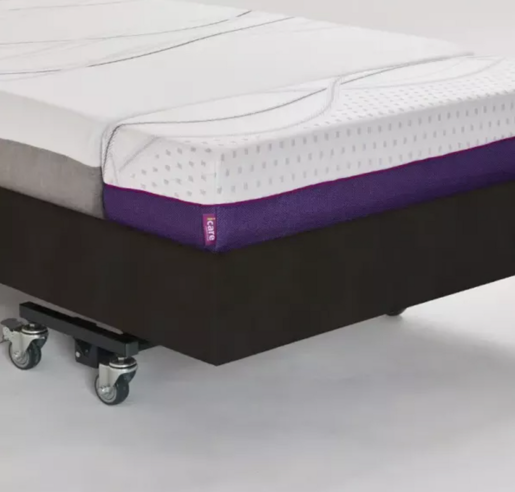 A mattress on a wheeled platform bed frame, shown in a studio setting. The mattress has a white textured top, a purple side bolster, and a gray side panel.